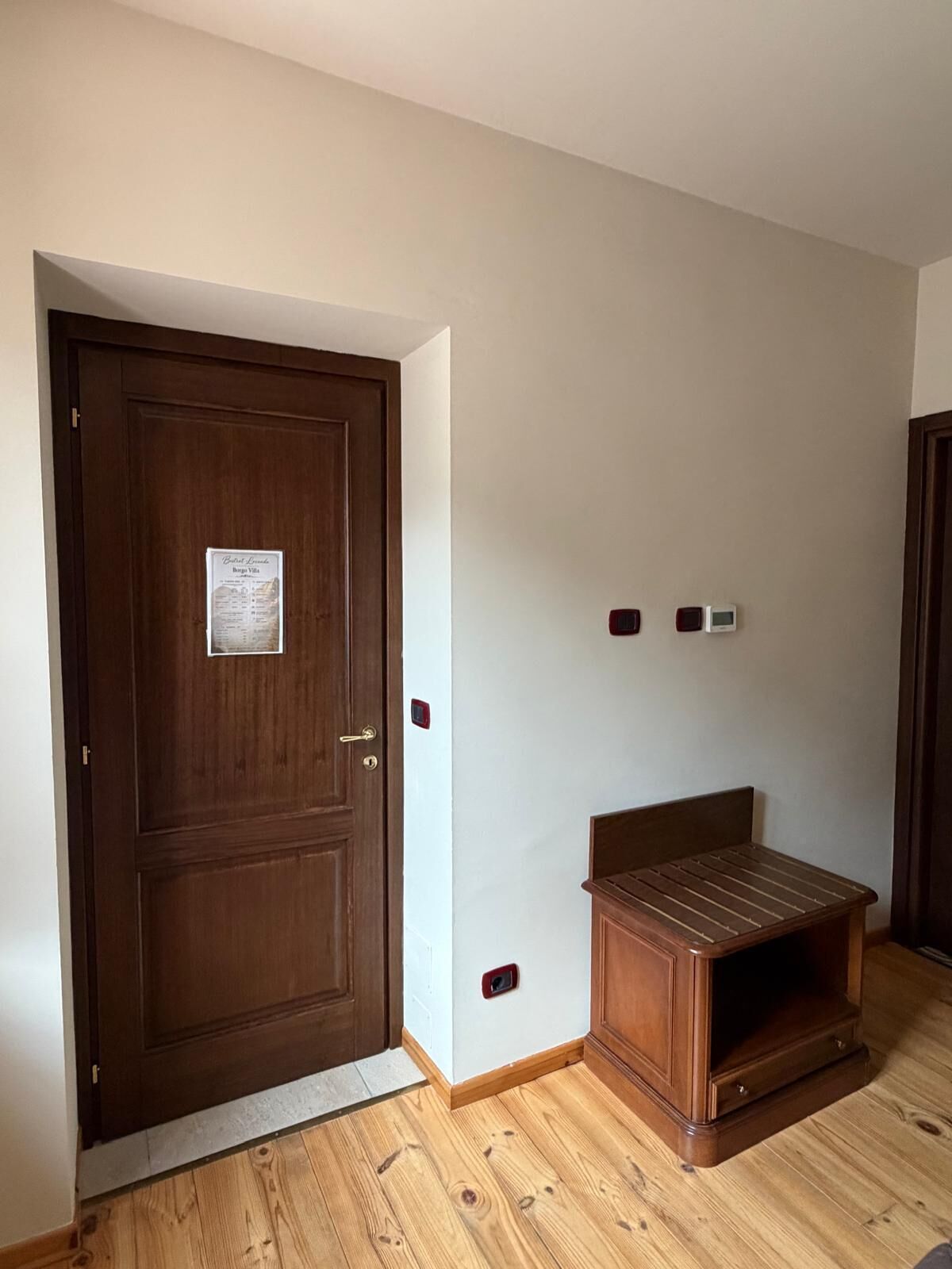 Una habitación con una puerta de madera, un pequeño banco de madera y un termostato en la pared.