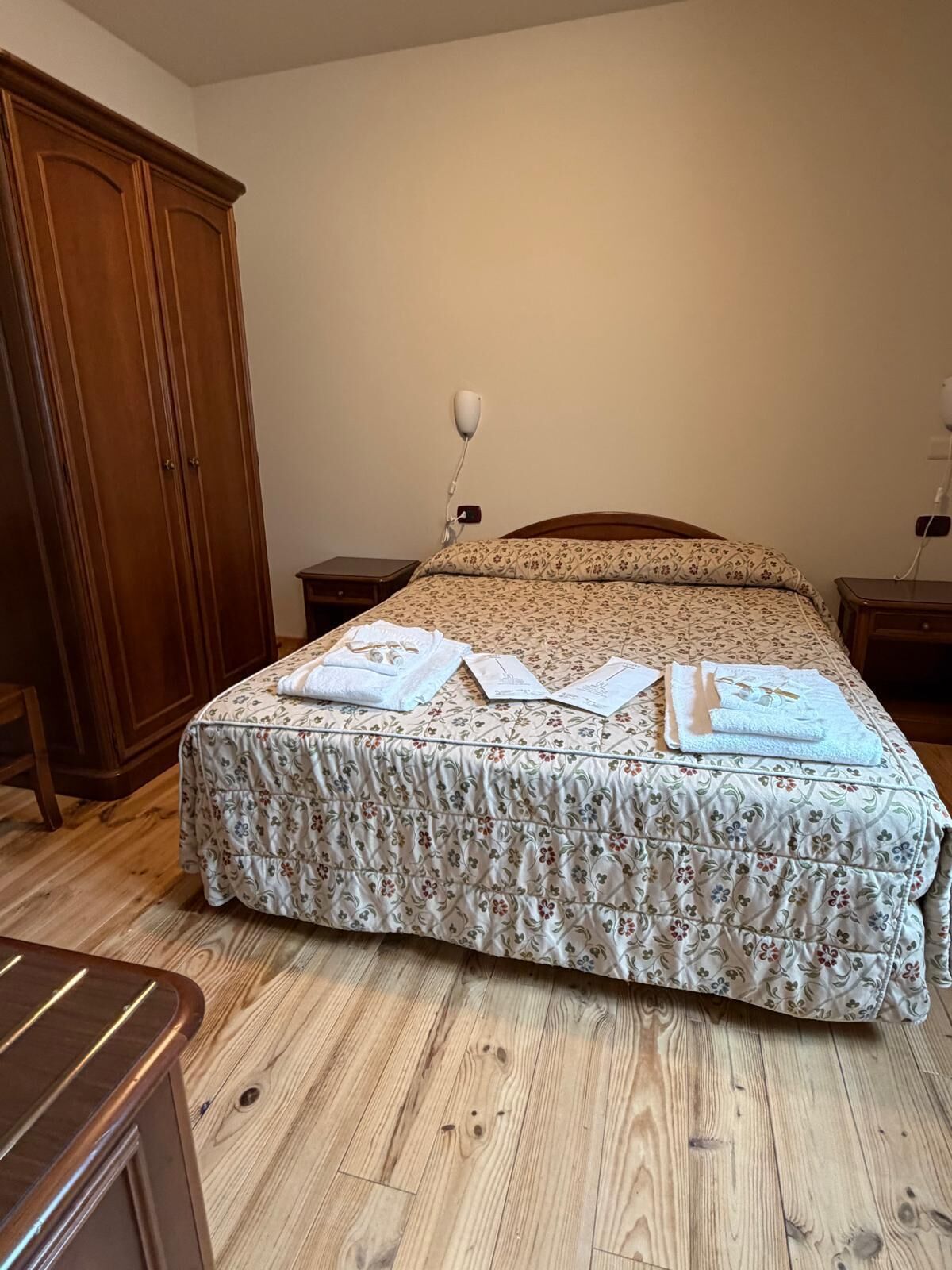 A bedroom with a wooden wardrobe, a bed with floral bedding, and a bedside table with a lamp.