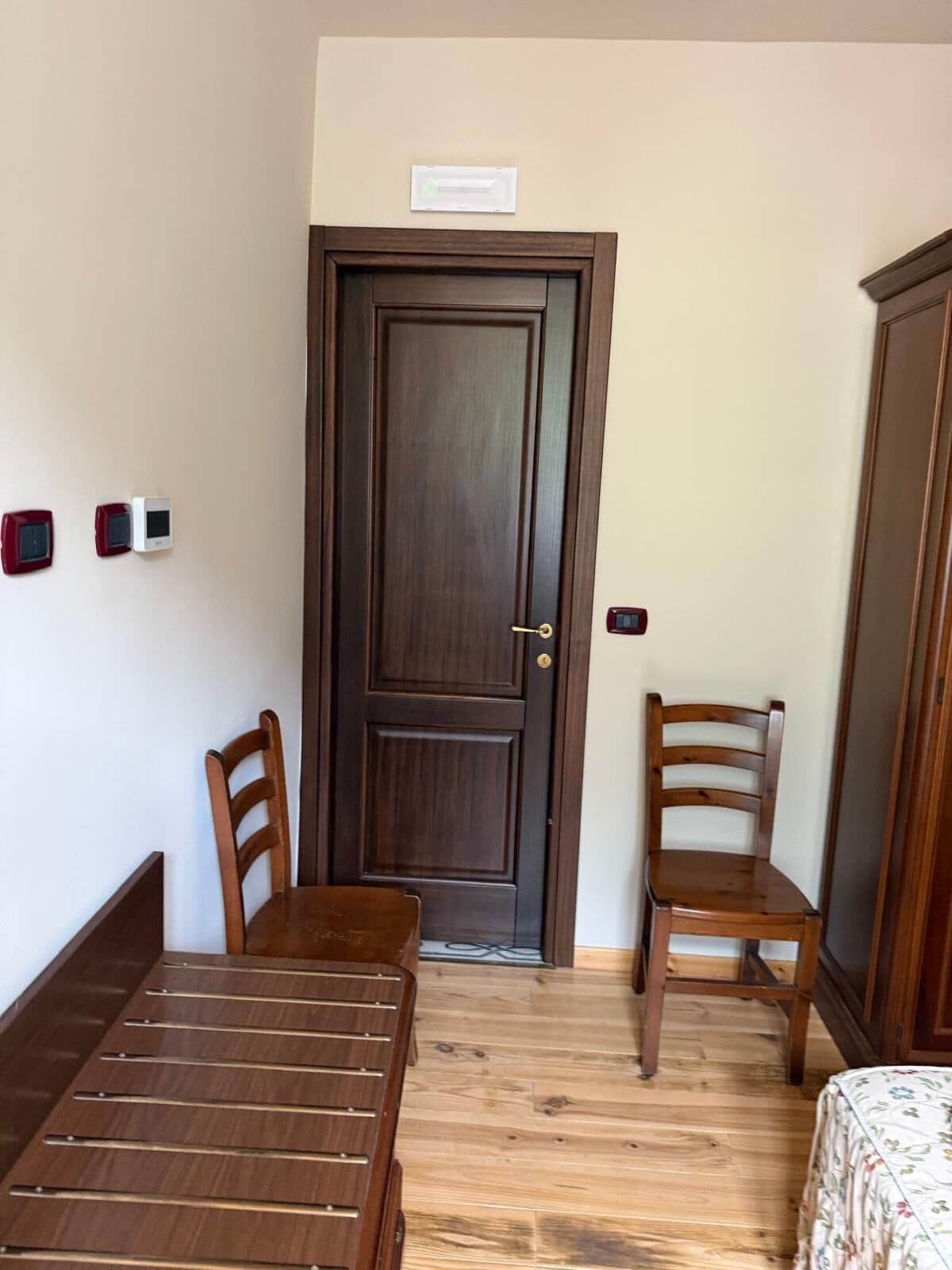 Une pièce avec une porte en bois, deux chaises en bois, une table en bois et un lit recouvert d’une courtepointe à motifs floraux.