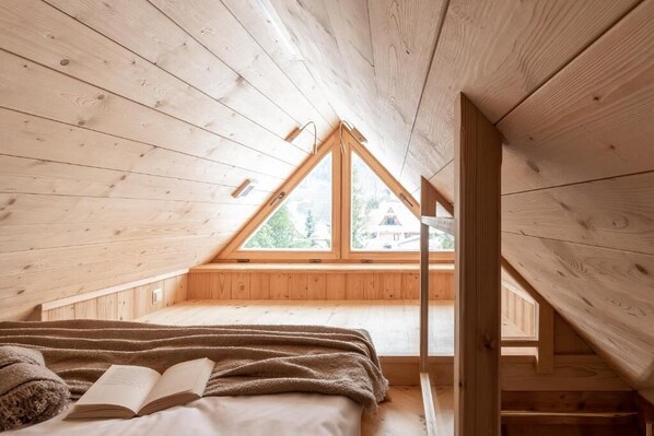 A wooden interior with a sloped ceiling, a bed with a blanket and book, and a window with a view of greenery.