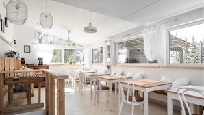 Una habitación luminosa y espaciosa con mesas de madera y sillas blancas, ventanas amplias y lámparas colgantes.