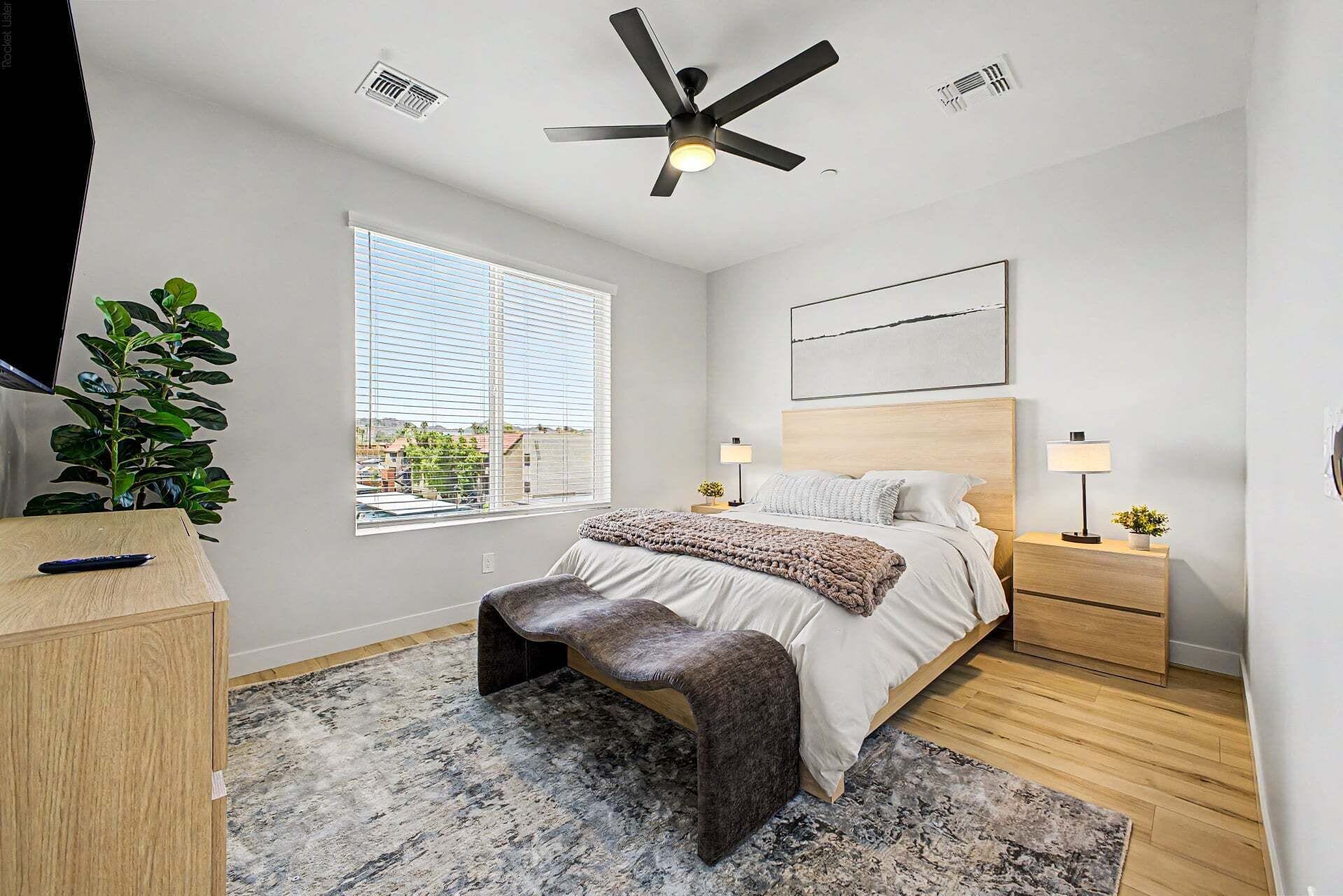 A bedroom with a bed, bedside tables, a ceiling fan, a window with blinds, a plant, and a desk with a chair.