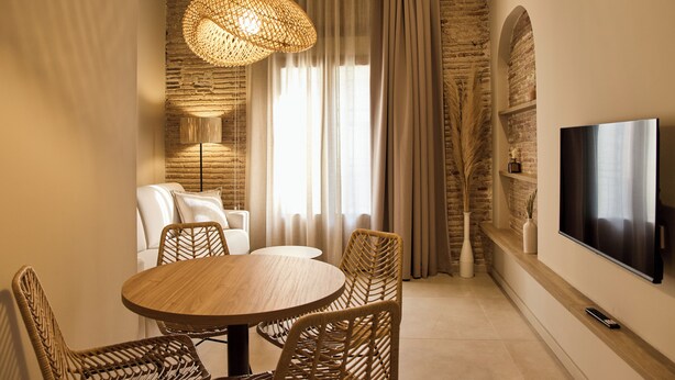 A modern living room with a round wooden table, wicker chairs, a flat-screen TV, and a large window with curtains. - Arrels Mercat (Valencia)