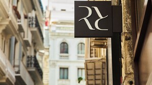 A narrow street with buildings on either side, a sign with a stylized 'R' logo, and a yellow awning. - Arrels Mercat (Valencia)