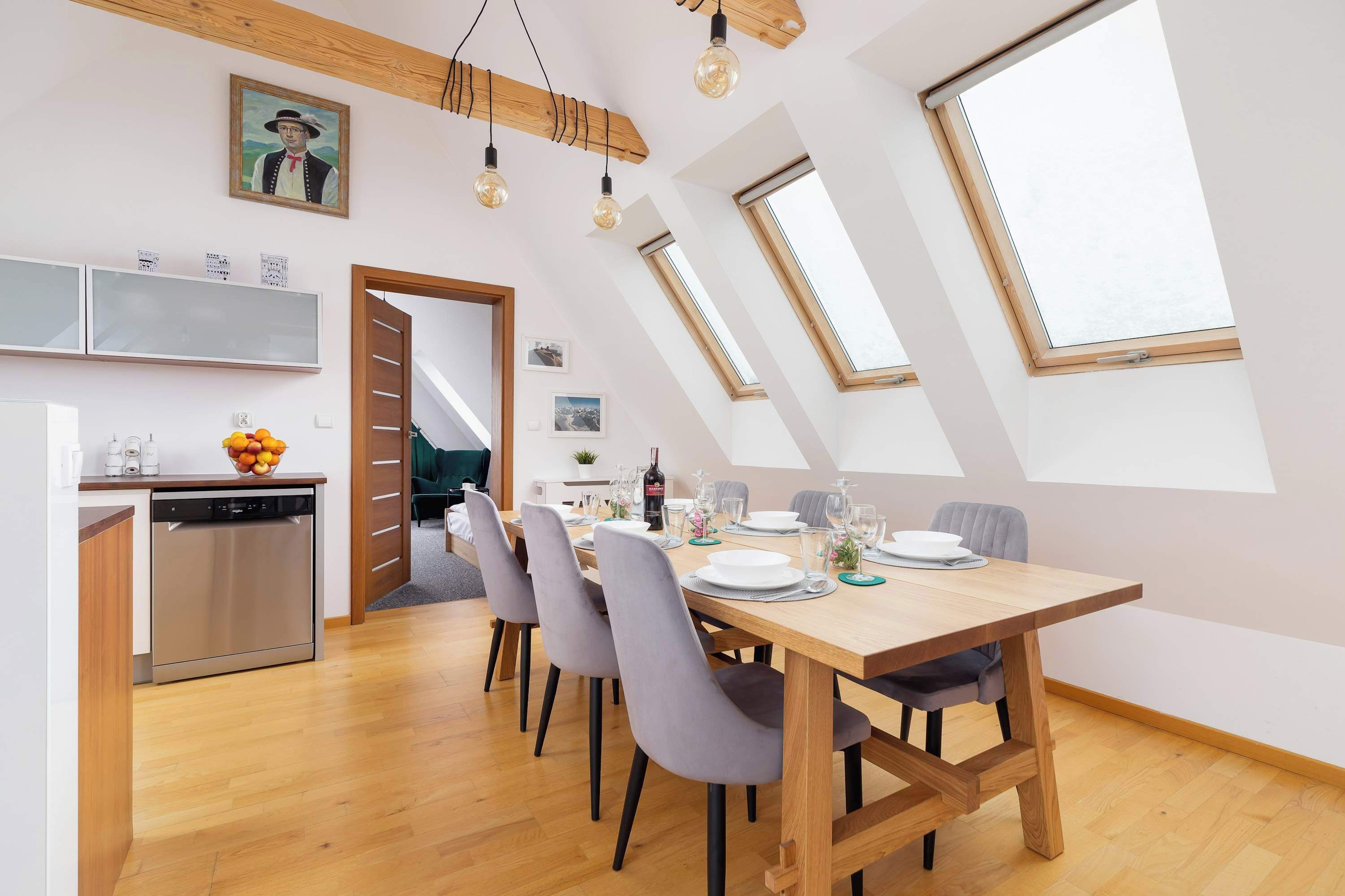 Bright open-plan dining area with a wooden dining table for four, large skylights, exposed wooden beams, and a modern kitchenette in the background.