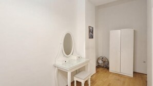 A white vanity with a mirror, a small drawer, and a bench. There is a white wardrobe and a fan on the floor. - Cozy 1BR Apt with Living Room & Garden (Vienna)