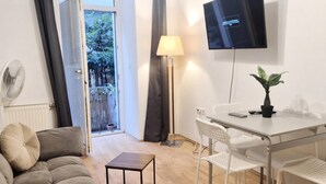 A hotel room with a sofa, a small table, a desk with chairs, a TV, a lamp, a potted plant, and a view through a glass door. - Cozy 1BR Apt with Living Room & Garden (Vienna)