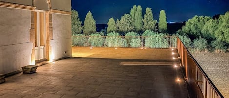 Un espacio al aire libre empedrado, con barandilla, entrada a un edificio y árboles de fondo.