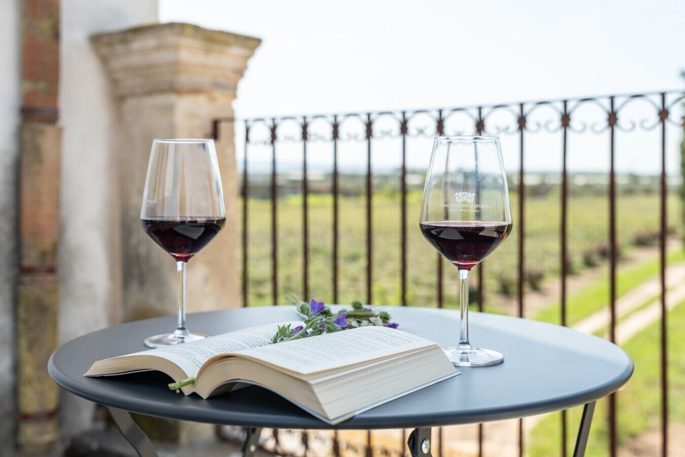 Une table avec deux verres à vin et un livre ouvert, placée devant la rambarde d’un balcon d’où l’on peut admirer la verdure.