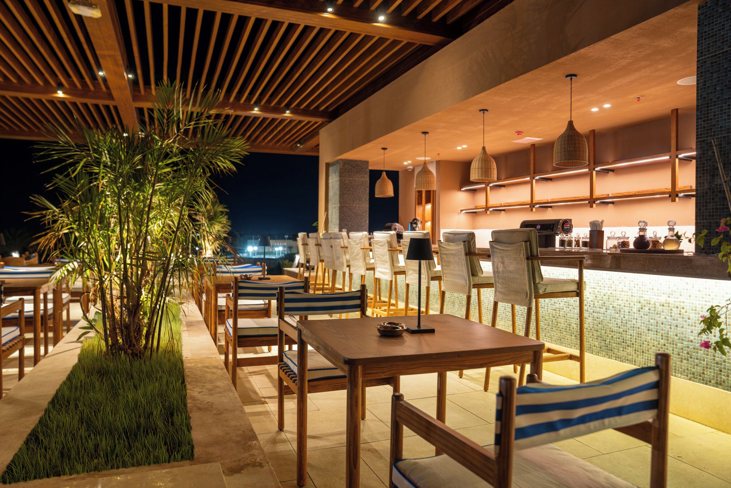 Uma área de refeições ao ar livre com mesas e cadeiras de madeira, um balcão de bar e luminárias pendentes.