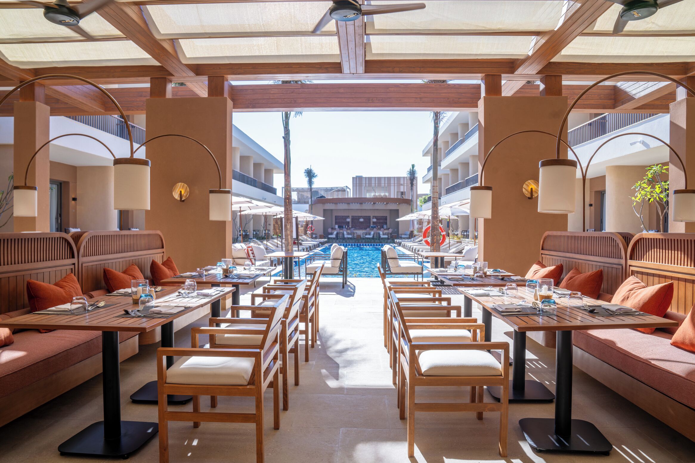 A sunlit outdoor dining area with tables and chairs, a pool, and a view of a building in the background.