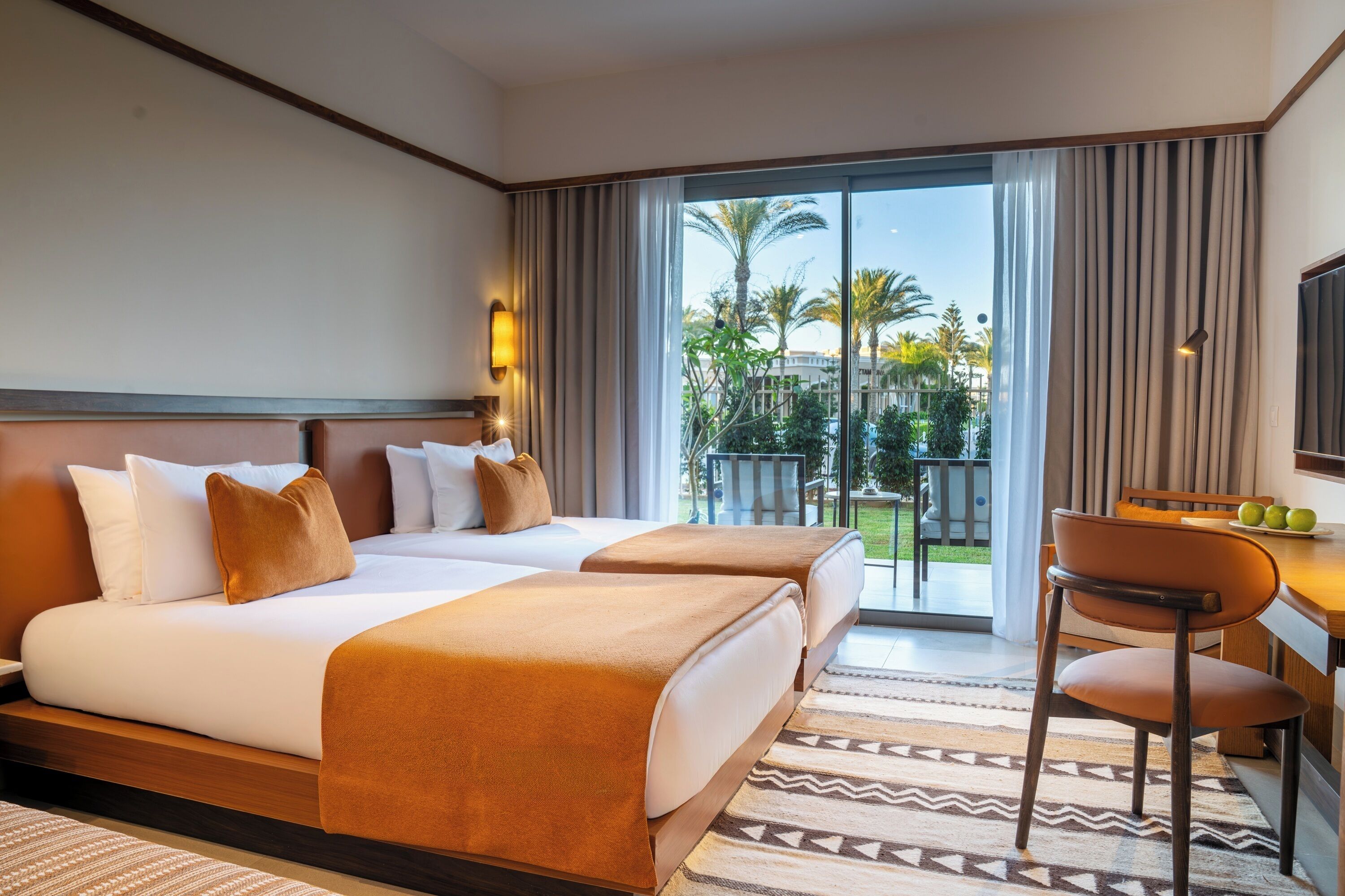 A hotel room with a bed, a desk, a chair, a TV, and a balcony with a view of palm trees.