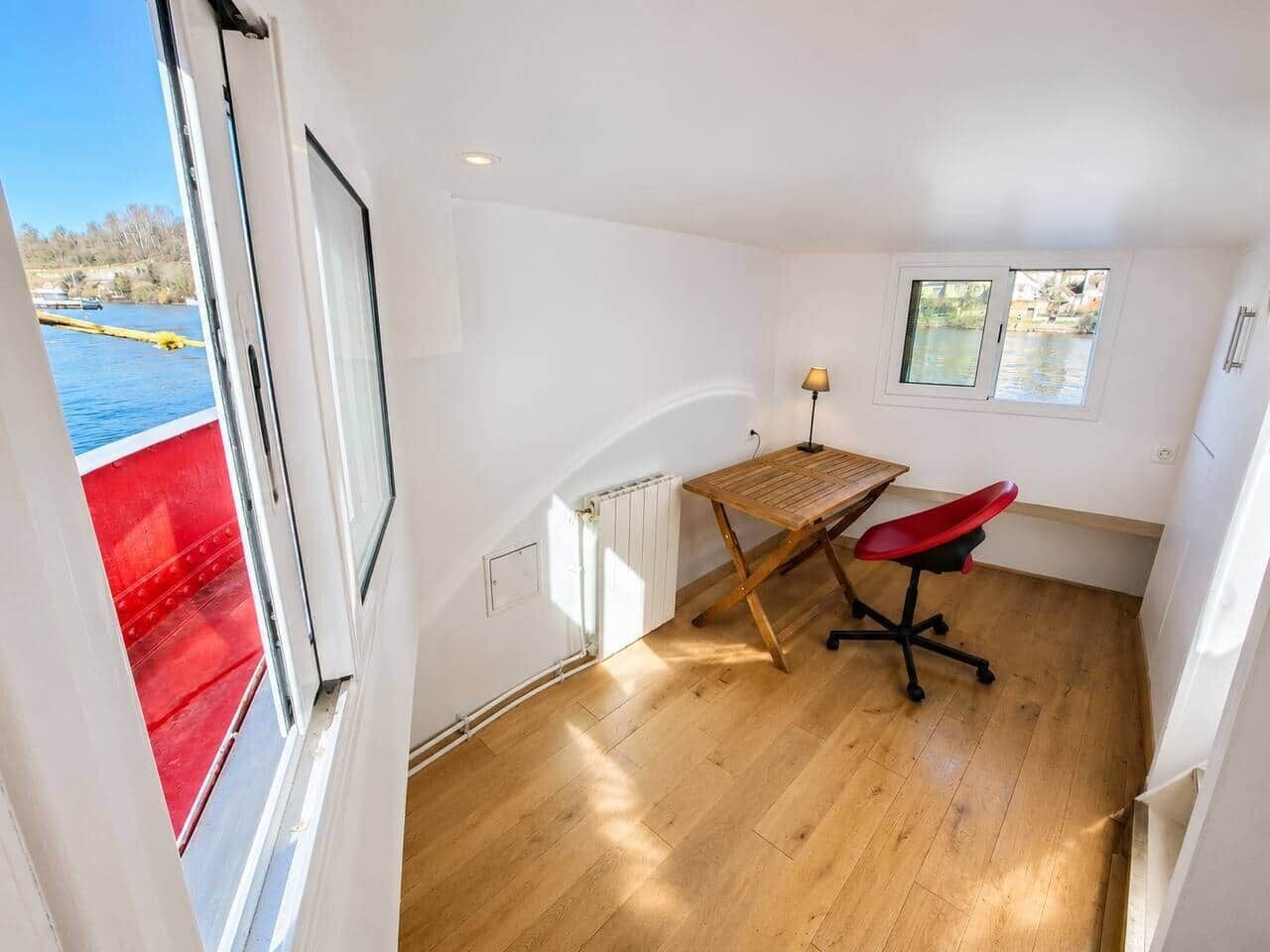 Een kamer met een houten vloer, een bureau met een rode stoel, een raam en uitzicht op een rode boot op het water.