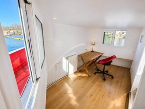 A room with a wooden floor, a desk with a red chair, a window, and a view of a red boat on the water.