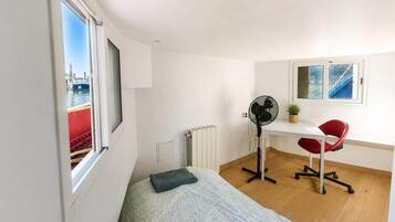 Een kamer met een bed, een bureau met stoel, een ventilator, een radiator en een raam met uitzicht op een boot.