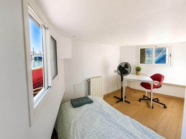 A room with a bed, a desk with a chair, a standing fan, a radiator, and a window with a view of a boat.