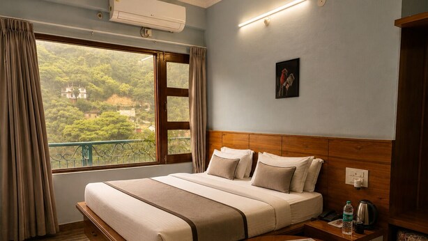 A hotel room with a bed, a round wooden table, a nightstand, a window with curtains, and an air conditioning unit. - Hotel Classio (Balichauki)