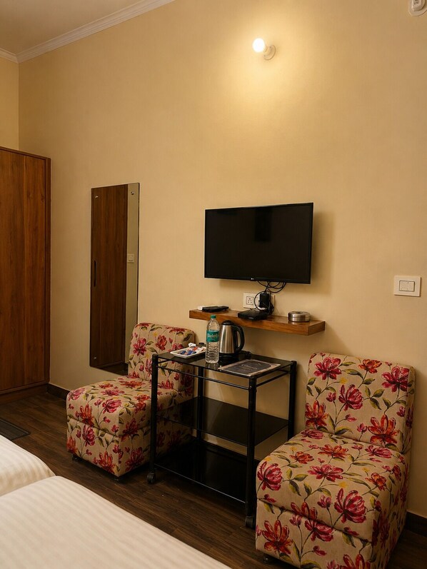 Une chambre d’hôtel avec un lit, deux fauteuils à motifs floraux, une petite table avec une bouteille et une bouilloire, un téléviseur fixé au mur et une armoire en bois.