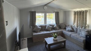 A modern living room with a sofa set, a coffee table with a plant, a television, and a window with curtains. - Static Caravan 3 Bed, Sleeps 6, Waterside, No Pets (Northampton)