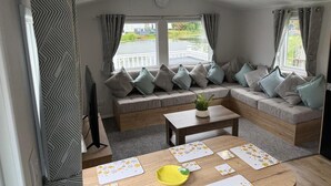 A modern interior with a sofa set, a dining table, and a view of a water body outside. - Static Caravan 3 Bed, Sleeps 6, Waterside, No Pets (Northampton)