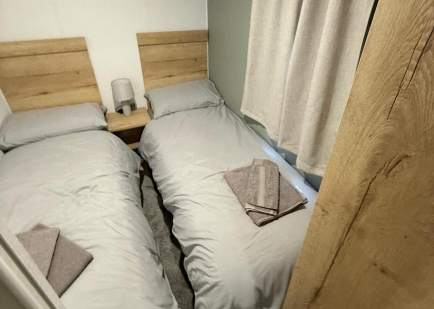 Un dormitorio con dos camas individuales, cabeceros de madera, una mesita de noche con una lámpara y una ventana con cortinas.