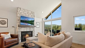 A living room with a stone fireplace, a leather sofa, a beige sofa, and a large window with a view of trees and hills. - Downtown WP Sojourn Villa 115 | Bunk Room | Great Views | Discounted Attractions (Winter Park)
