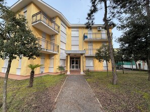 A multi-story building with balconies, a central entrance, and a surrounding grassy area with trees. - Great flat with two terraces and parking (Bibione)