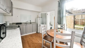 A modern kitchen with grey cabinets, a wooden dining table, and a view of a brick garden. - Host My Nest: 2-Bed w/ Garden Near Forest Gate (london)