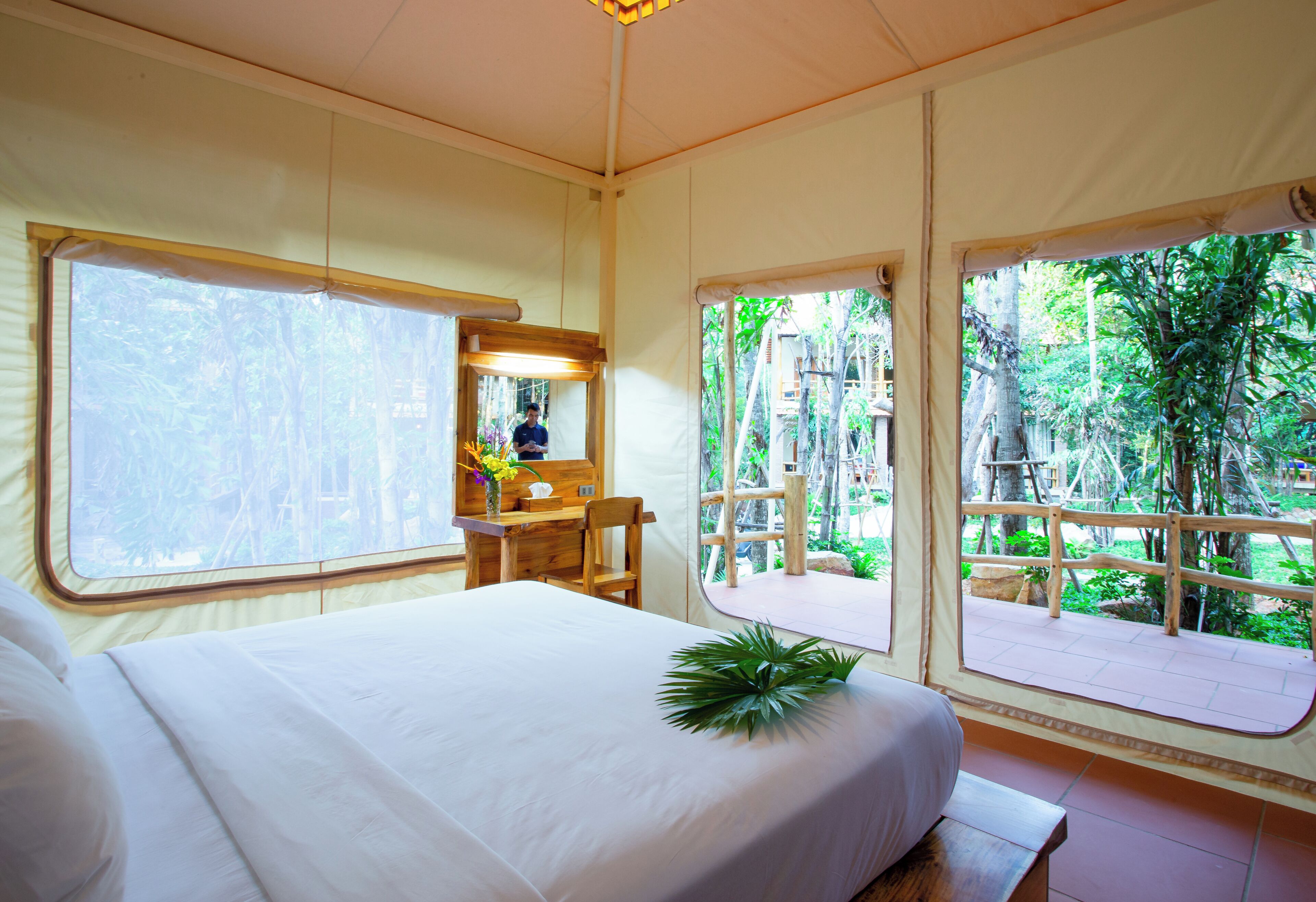 A room with a bed, a desk, and large windows offering a view of a garden.