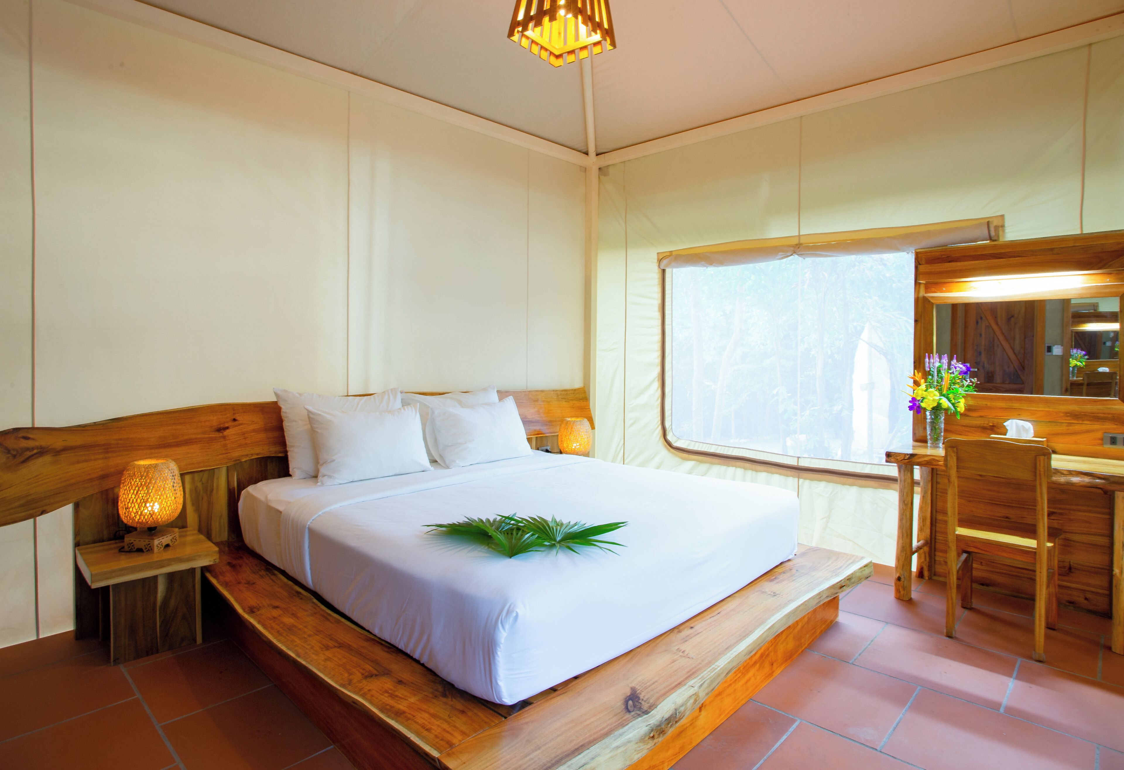 A bedroom with a wooden bed, white bedding, a bedside lamp, a desk with a mirror, and a window with a view of trees.