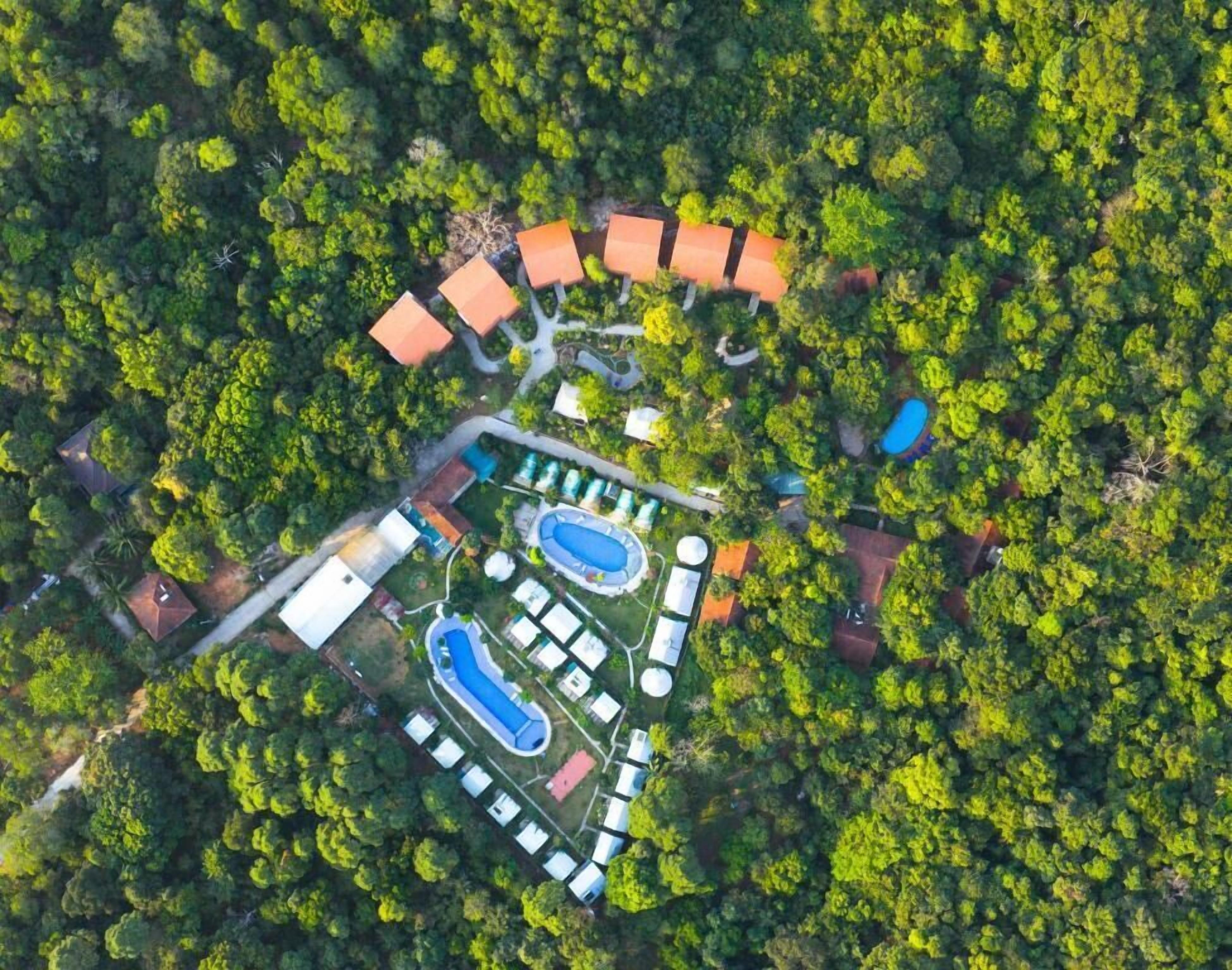 An aerial view of a resort with a swimming pool, tennis courts, and a cluster of buildings surrounded by dense forest.