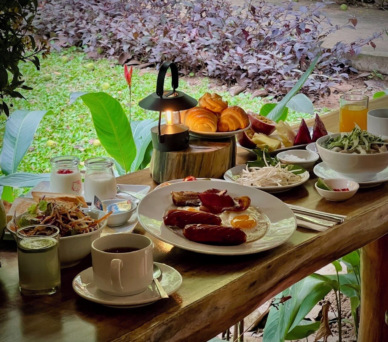 A wooden table set with breakfast items including eggs, sausages, and pastries, with a view of a garden in the background.