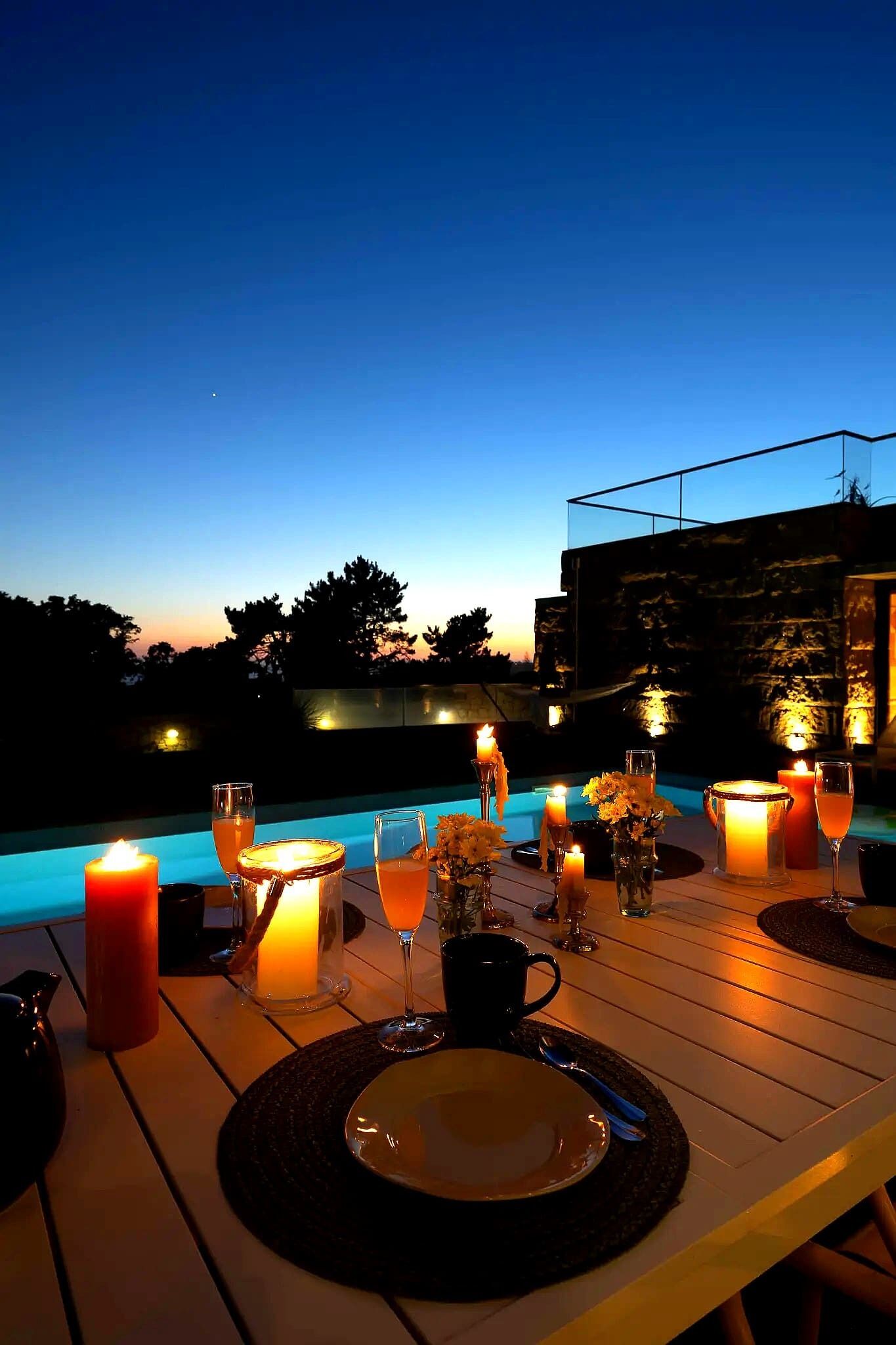 Un área de comedor al aire libre con una mesa dispuesta para dos personas, velas y vistas a una piscina al atardecer.