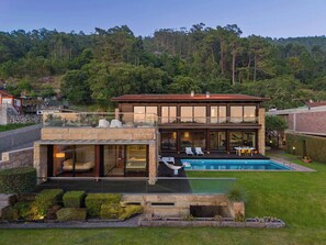 Ein modernes, zweistöckiges Haus mit Swimmingpool, Natursteinaußenwand und Blick auf einen bewaldeten Hügel.