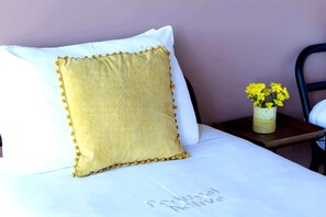 Un lit avec un oreiller blanc et un oreiller jaune texturé agrémenté de glands. Un vase contenant des fleurs jaunes est posé sur une table de chevet.