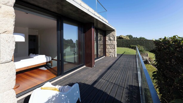 A modern outdoor deck with a wooden floor, a glass door leading to a room with a bed and a white chair, and a clear view of a landscaped garden. - Sunrise Escape with Pool by Portugal Active (Viana do Castelo)