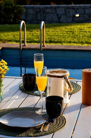 Una mesa de comedor dispuesta con platos, cubiertos, vasos y una jarra de jugo, con una piscina y follaje al fondo.