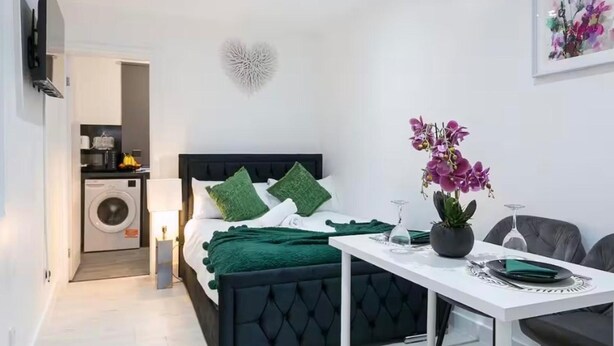 A modern bedroom with a black headboard, green pillows, a white bedside table, a potted orchid, and a dining area with a chair and table.