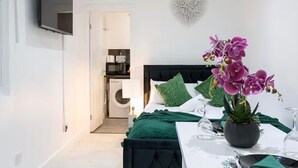 A bedroom with a bed, green pillows, a potted orchid, a bedside table with a glass, and a TV mounted on the wall.