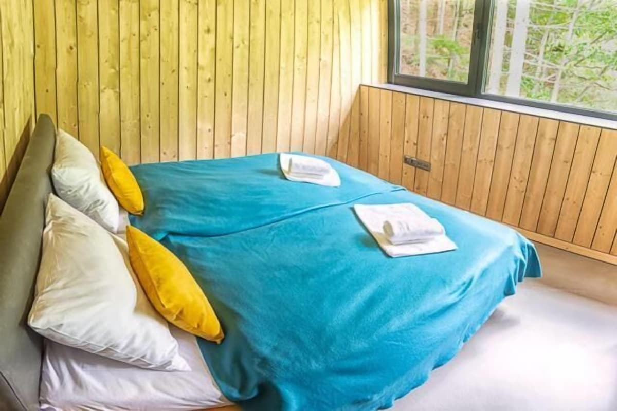 Una cama con colcha y almohadas azules, una pared de madera y una ventana con vistas.