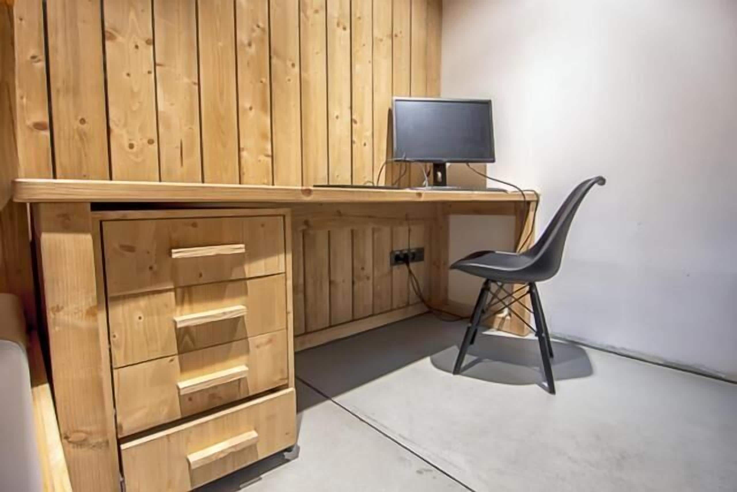 A wooden desk with drawers and a computer monitor, a black chair, and a concrete floor.