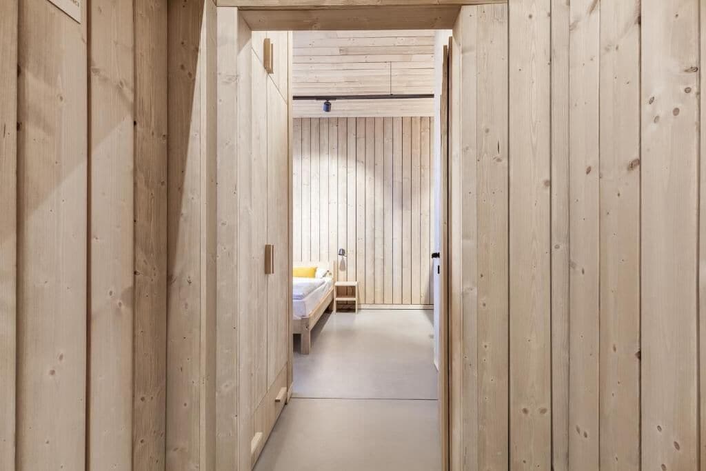 A narrow hallway with wooden walls leading to a bedroom with a bed and a nightstand.