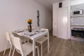 A dining area with a white table set for four, a vase with yellow flowers, and a modern kitchen with a microwave and oven. - Castle Harbour 4 - 1-Bedroom Apartment (Los Cristianos)