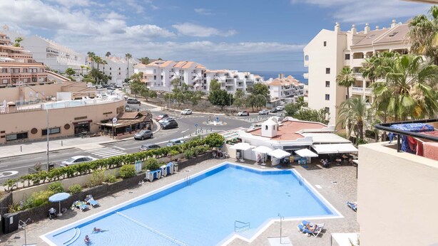 A pool area with lounge chairs, a street view with buildings, and a clear sky. - Castle Harbour 4 - 1-Bedroom Apartment (Los Cristianos)