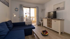 A living room with a sofa, a television on a cabinet, a dining area with a table and chairs, and a view through a glass door. - Castle Harbour 4 - 1-Bedroom Apartment (Los Cristianos)
