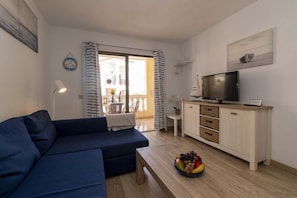 A living room with a sofa, a television on a cabinet, a dining area with a table and chairs, and a view through a glass door. - Castle Harbour 4 - 1-Bedroom Apartment (Los Cristianos)