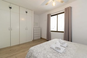 A bedroom with a bed, a wardrobe, a dresser, and a window with curtains. - Castle Harbour 4 - 1-Bedroom Apartment (Los Cristianos)