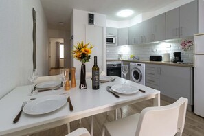 A modern kitchen with white cabinets, a dining table set with plates, wine glasses, and a bottle. - Castle Harbour 4 - 1-Bedroom Apartment (Los Cristianos)
