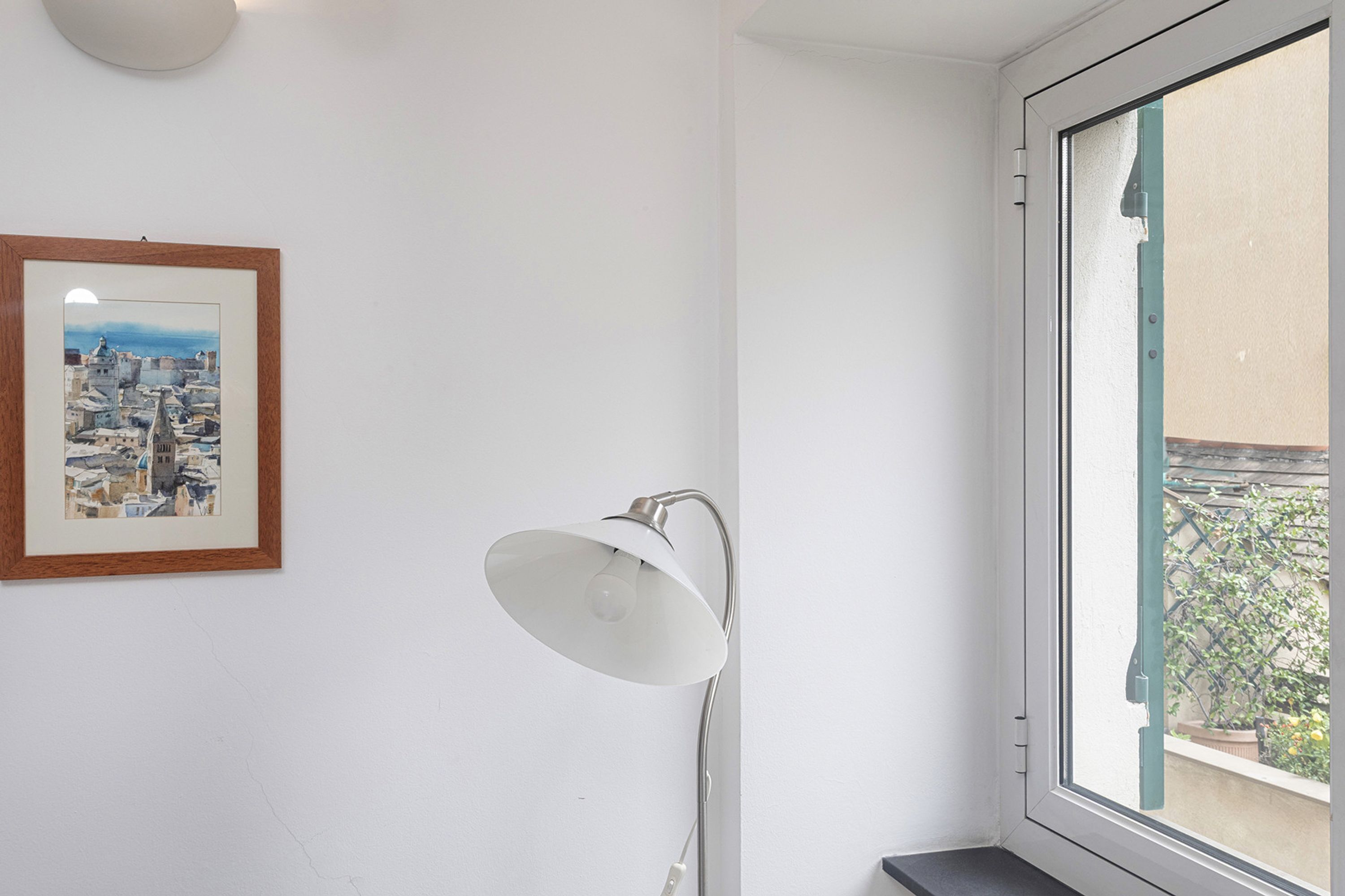 Een kamer met een stadsgezicht in een lijst, een vloerlamp en een raam met uitzicht op een gebouw en groen.