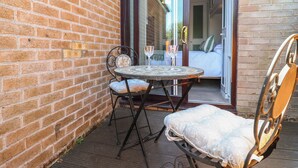 Un patio con una mesa redonda y dos sillas, un muro de ladrillo y un ventanal corredero que da a un dormitorio.